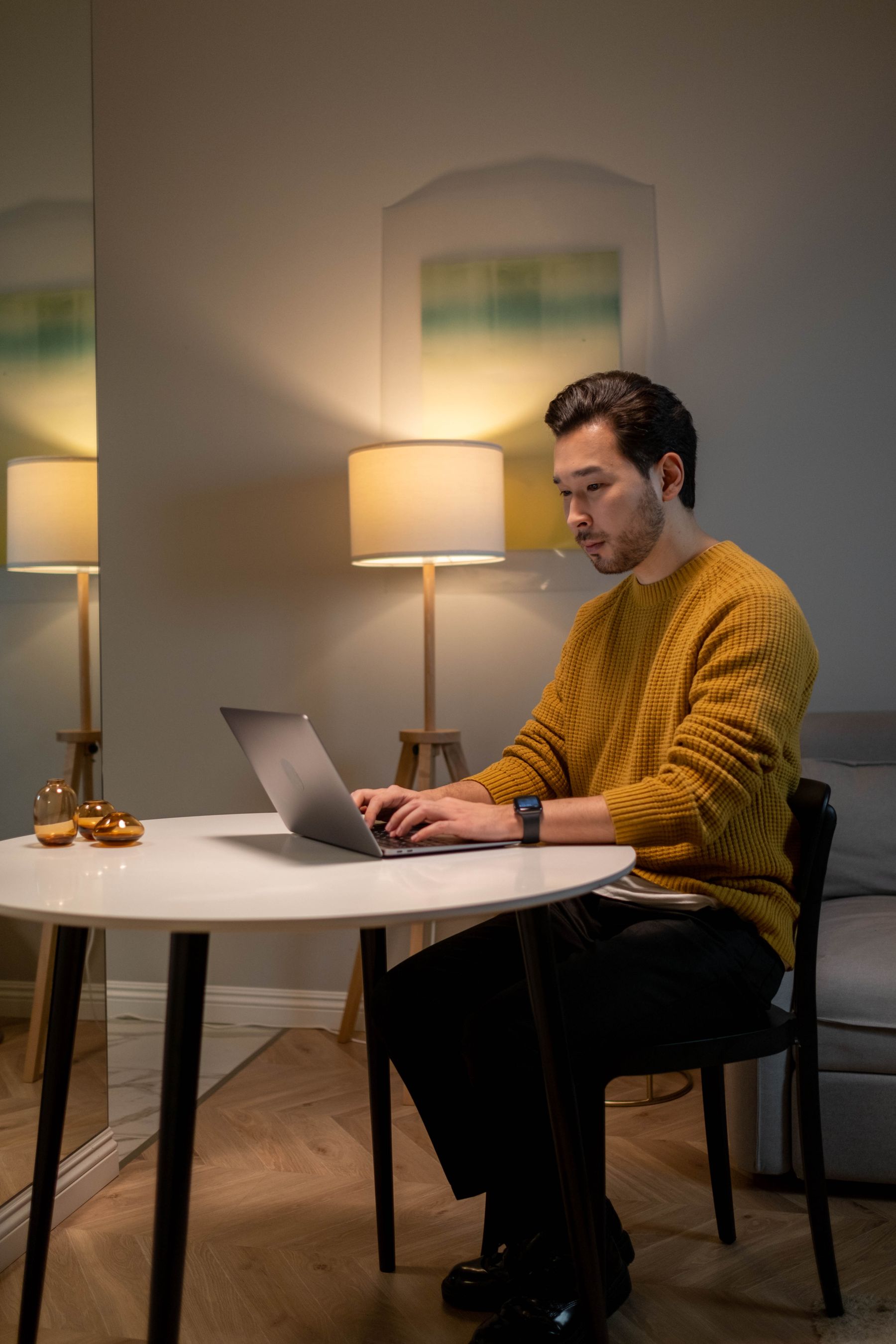 A designer working late at a warmly lit desk, laptop open, reflected in a mirror.